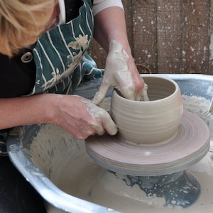May include: A potter at work, using a pottery wheel to form a light-coloured clay bowl. The potter's hands are covered in wet clay as they shape the vessel. The wheel is spinning, and the bowl is taking form. The potter is wearing a green and white striped apron.