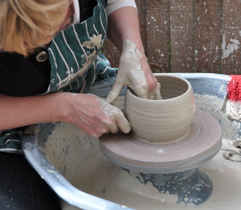 May include: A potter at work, using a pottery wheel to form a light-coloured clay bowl. The potter's hands are covered in wet clay as they shape the vessel. The wheel is spinning, and the bowl is taking form. The potter is wearing a green and white striped apron.
