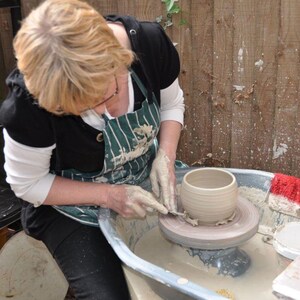 May include: A person shaping a light brown ceramic pot on a pottery wheel. The potter wears a green and white striped apron and has clay-covered hands. The pot has a rounded shape with horizontal lines. The background features a wooden fence.