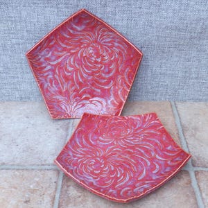 Puede incluir: Dos platos de cerámica rojos con un estampado floral. Los platos tienen forma pentagonal con un borde elevado. El diseño floral es en un tono más claro de rojo y blanco, creando contraste. Los platos están sobre una superficie embaldosada.