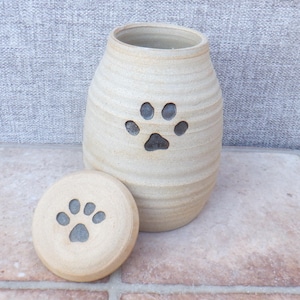 Puede incluir: Urna para mascotas de cerámica beige con diseño de huella de pata. La urna tiene forma cilíndrica con tapa, y las huellas de pata están talladas en la superficie. Diseñada para contener las cenizas de una mascota.