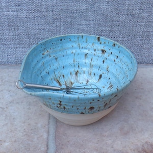 Puede incluir: Un cuenco mezclador de cerámica hecho a mano con interior azul claro y base color crema. El cuenco tiene un pico vertedor y está moteado con marrón. Un pequeño batidor de acero inoxidable descansa dentro. Utensilios de cocina.