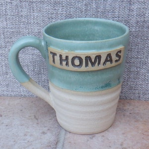 Puede incluir: Una taza de cerámica azul claro y blanco con una franja marrón y el nombre "THOMAS" escrito en ella.