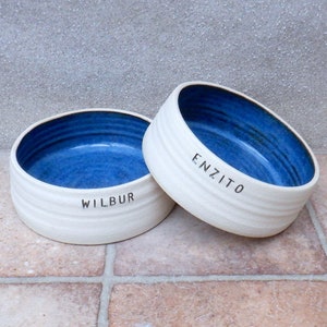 May include: Two white ceramic pet bowls with blue interiors. The bowls are personalised with the names "Wilbur" and "Enzito".
