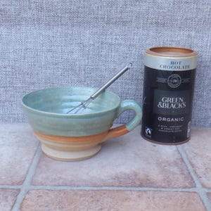 May include: A ceramic mug with a light blue glazed interior and upper exterior, transitioning to a natural tan base. A metal whisk rests inside the mug. Beside it stands a cylindrical black container labeled Green & Black's Organic Hot Chocolate.
