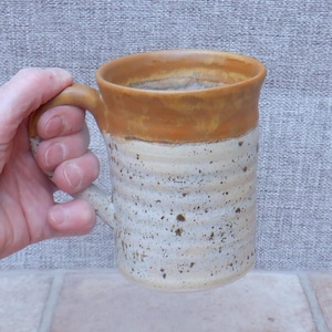 Könnte beinhalten: Ein Keramikbecher mit zylindrischem Körper, horizontalen Rillen und einer gesprenkelten, cremeweißen Glasur. Der obere Rand und der Henkel sind mit einer kontrastierenden, bernsteinfarbenen Glasur versehen, was dem Gefäß eine zweifarbige Optik verleiht.
