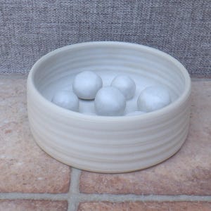 Puede incluir: Un cuenco redondo de cer&aacute;mica de color blanco roto con una textura exterior acanalada. El interior del cuenco contiene varias bolas de cer&aacute;mica lisas, esf&eacute;ricas y de color blanco roto de distintos tama&ntilde;os, creando una composici&oacute;n sencilla.