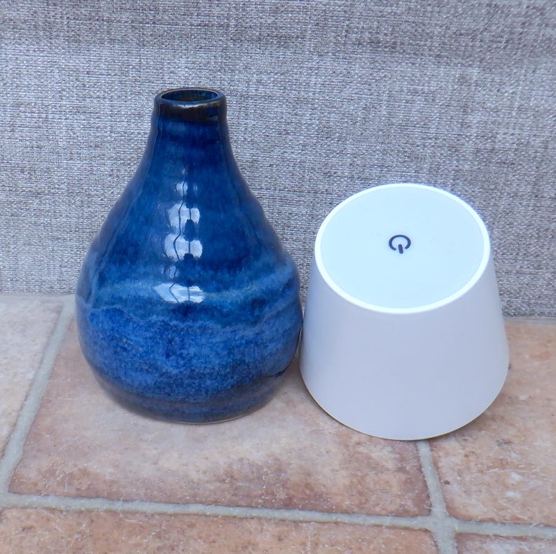May include: A glossy, deep blue ceramic vase with a tapered neck and a rounded body, positioned next to a white, conical electronic device featuring a circular power button symbol on its flat top surface, both resting on a tiled surface.