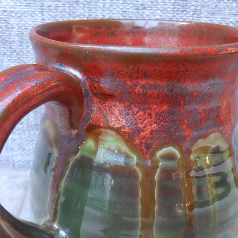 May include: A handmade ceramic mug with a large handle. The mug features a vibrant colour scheme, with a deep red rim and handle, transitioning to shades of green and brown on the body. The glaze has a glossy finish.