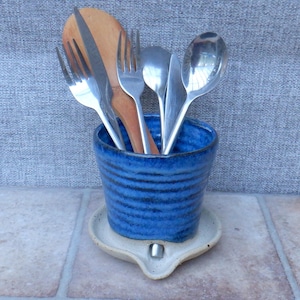 Könnte beinhalten: Ein blauer Keramik-Utensilienhalter mit passendem Löffelhalter. Der Halter enthält einen Holzlöffel, ein Messer, Gabeln und Löffel. Der Halter hat horizontale Linien und steht auf einer cremefarbenen Löffelablage.