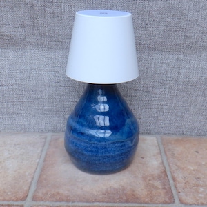 Peut inclure: Une lampe de table composée d'une base en céramique bleu profond et brillant, de forme arrondie et bulbeuse, surmontée d'un abat-jour conique blanc uni. La base bleue présente une surface réfléchissante avec des reflets lumineux visibles.