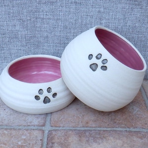 Puede incluir: Dos cuencos de cerámica blanca para mascotas con interiores rosados. Cada cuenco tiene un diseño de huella de pata gris.