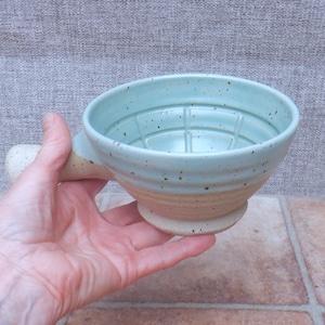 Puede incluir: Un cuenco de afeitar de cerámica con interior esmaltado en azul claro que presenta un patrón de rejilla en la base. El exterior tiene un acabado moteado con una base de arcilla color canela natural y un mango corto y resistente.
