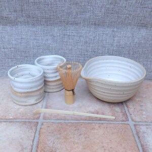 May include: A matcha tea set consisting of two small ceramic cups, one large ceramic bowl, a bamboo whisk, and a bamboo scoop. The ceramic pieces feature a white glazed interior with a speckled finish and a ribbed, textured exterior surface.