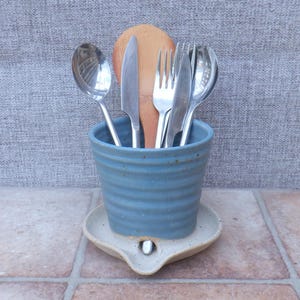 May include: A blue ceramic utensil holder with horizontal ridges, resting on a matching light-colored ceramic base. The holder contains a wooden spoon, two stainless steel knives, two stainless steel forks, and a stainless steel spoon.