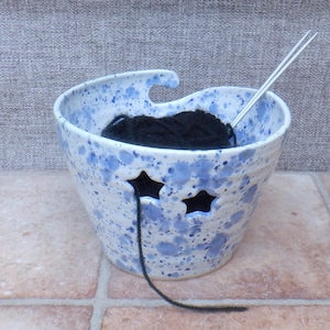 May include: A ceramic yarn bowl with a speckled blue and white design. The bowl has two star-shaped cutouts and a curved handle. Black yarn is inside the bowl, with two silver knitting needles and a strand of yarn extending from the star cutouts.