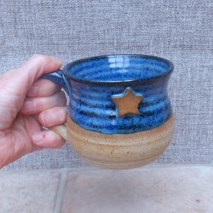 May include: A ceramic mug featuring a two-tone design with a glossy deep blue upper section and a natural tan lower section. A small, raised, five-pointed tan star is centered on the blue portion. The mug has a curved handle and a wide rim.