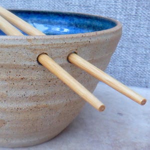 May include: A handcrafted ceramic bowl with a speckled tan exterior and a bright blue interior. Two wooden chopsticks are inserted through the bowl's side. The bowl is designed for serving Asian-inspired dishes.