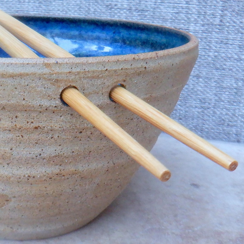 May include: A handcrafted ceramic bowl with a speckled tan exterior and a bright blue interior. Two wooden chopsticks are inserted through the bowl's side. The bowl is designed for serving Asian-inspired dishes.