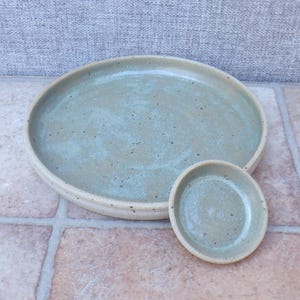 Może przedstawiać: Ręcznie wykonany zestaw ceramiczny talerz i mała miseczka. Talerz jest duży i okrągły, z jasnozieloną, nakrapianą glazurą i naturalnym, nieszkliwionym brzegiem. Mniejsza miska odzwierciedla wzór talerza.
