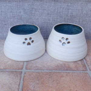 May include: Two white ceramic pet bowls with a blue glaze inside and a paw print design. The bowls are on a tiled floor.