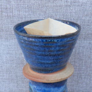 Puede incluir: Un gotero de café de cerámica con un acabado esmaltado en azul profundo, estrías horizontales y una base natural sin esmaltar de color tostado. Un filtro de café de papel beige claro está insertado en la abertura superior del cono.