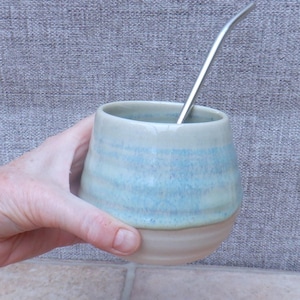 Puede incluir: Una taza de cerámica azul claro y beige con una pajita de metal. La taza tiene un fondo redondeado y una abertura ancha.