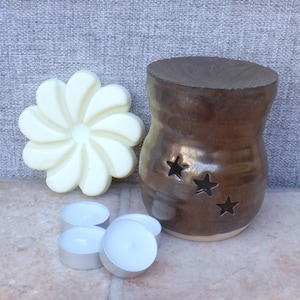May include: A brown ceramic wax warmer with three star cutouts. The warmer is on a beige surface with four white tea lights and a white wax melt in the shape of a flower.