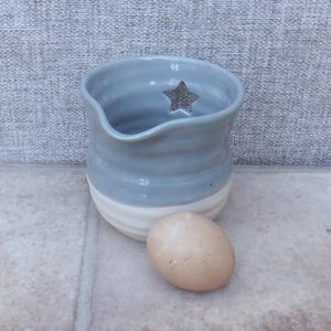 May include: A handmade ceramic pitcher with a light blue and white glaze. The pitcher has a star-shaped cutout and is next to a brown egg. The pitcher is approximately 4 inches tall.