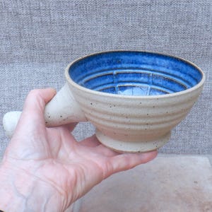 May include: Handmade ceramic shaving bowl with a blue interior and a speckled, beige exterior. The bowl has a handle and a footed base. The interior features a grid pattern. The bowl is held in a hand.