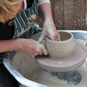 May include: Hands shaping a light-coloured clay pot on a pottery wheel. The hands are covered in wet clay, and the wheel is spinning. The person wears a green and white striped apron. The pottery wheel is in a metal basin.