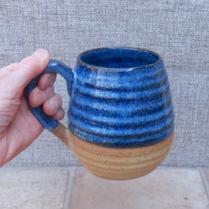 May include: A ceramic mug featuring a two-tone design with a glossy, deep blue glazed upper section and a natural, unglazed tan base. The mug has a rounded, bulbous shape with horizontal ribbed textures throughout the body and a curved handle.