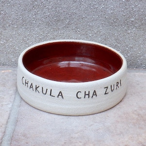 Puede incluir: Tazón de cerámica blanco para mascotas con interior rojo. El tazón tiene las palabras "Chakula Cha Zuri" inscritas en el exterior.
