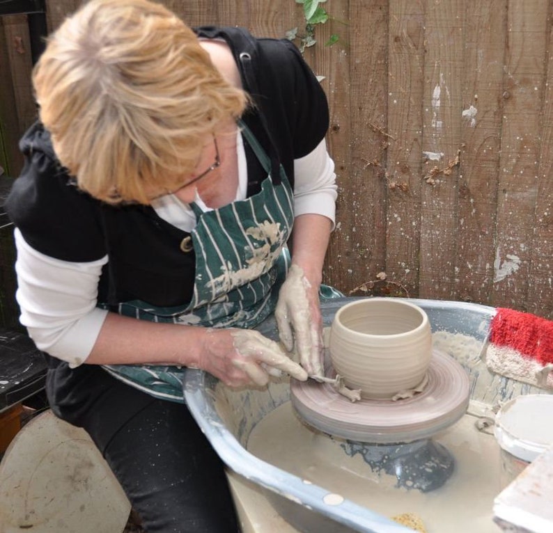 Puede incluir: Una vasija de arcilla beige siendo moldeada en un torno de alfarero. El recipiente tiene un cuerpo redondeado y una abertura ancha. El torno est&aacute; dentro de una cubeta con residuos de arcilla h&uacute;meda y una peque&ntilde;a esponja roja.