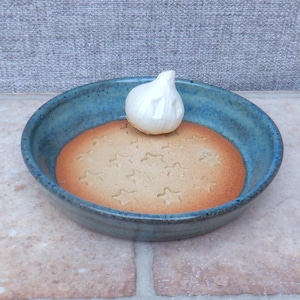 May include: A handmade, round, blue ceramic bowl with a light brown, star-patterned interior. A whole garlic bulb sits in the center. The bowl has a rustic, artisan appearance, suitable for serving or display.