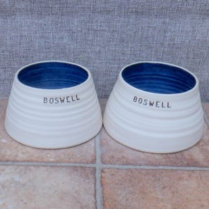 May include: Two white ceramic bowls with a blue interior and the word "BOSWELL" printed on the side. The bowls have a ridged texture and a flared shape, suitable for serving food or as decorative items.