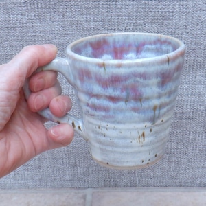 Op de afbeelding: Een handgemaakte keramische mok met een unieke glazuur. De mok heeft een lichtgekleurde basis met horizontale banden in roze en blauwe tinten. De handgreep is in het ontwerp geïntegreerd.