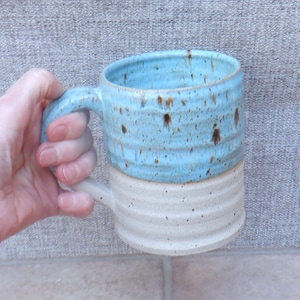 Könnte beinhalten: Ein Keramikbecher mit zweifarbigem Design, bestehend aus einem hellblauen oberen Teil und einem cremefarbenen unteren Teil. Der Becher hat eine gerippte, strukturierte Oberfläche und ist sowohl im blauen als auch im cremefarbenen Bereich gesprenkelt.