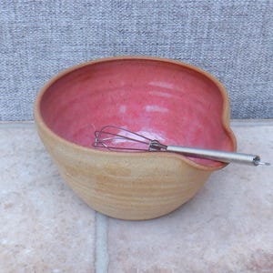 Könnte beinhalten: Eine handgefertigte Keramik-Rührschüssel mit leuchtend rosa Innenraum und natürlicher, unglasierter Außenseite. Ein Schneebesen aus Edelstahl liegt in der Schüssel. Die Schüssel hat eine einzigartige, asymmetrische Form, für die Küche.
