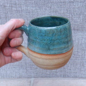 May include: A handmade ceramic mug with a teal blue glazed top and a tan base. The mug has a rounded shape with a handle and is held in a hand. The mug is approximately 4 inches tall.