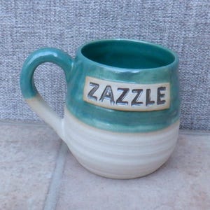 May include: A handmade ceramic mug in teal and cream. The word "ZAZZLE" is embossed on the teal section. The mug has a rounded shape and a large handle. A perfect gift for tea or coffee lovers.