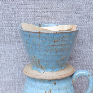 May include: A ceramic coffee pour-over dripper with a light blue glaze and brown speckled accents. The dripper has a wide, circular base that rests on a matching mug, and it contains a folded brown paper coffee filter inside the basin.
