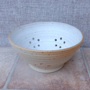 May include: A handmade ceramic colander with a cream-colored interior and a speckled, tan exterior. The bowl has several drainage holes and sits on a small, round base. The colander is designed for washing and draining fruits and vegetables.