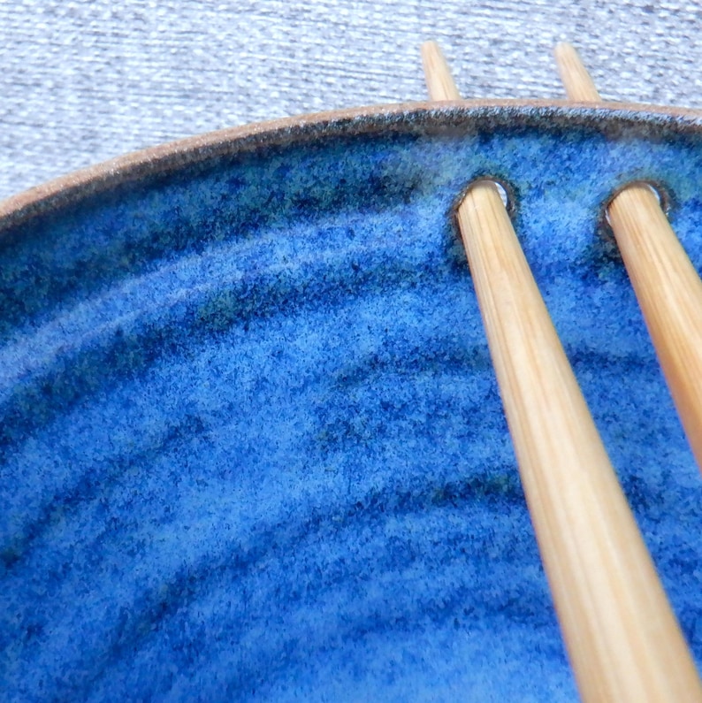 May include: A close-up shot of a handmade ceramic bowl with a vibrant blue glaze and a textured surface. Two wooden chopsticks rest in the bowl's designated slots. The rim of the bowl is brown.