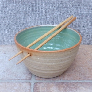 May include: A round ceramic bowl with a textured, light tan exterior and a smooth, speckled mint green interior. The bowl features two circular holes near the rim, through which a pair of light-colored wooden chopsticks are inserted.
