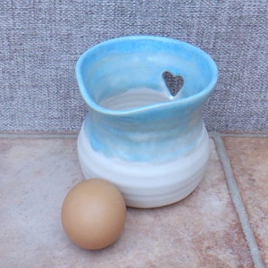 Puede incluir: Un porta huevos de cerámica azul claro con un recorte en forma de corazón. El porta huevos está esmaltado con un efecto degradado blanco y azul. Un huevo marrón está sentado junto al porta huevos.