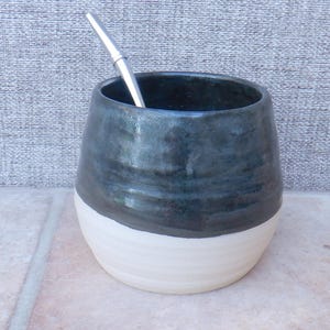 May include: A rounded ceramic mate cup featuring a two-tone design with a glossy dark charcoal upper section and a matte off-white base. A slender, polished silver-colored metal bombilla straw is inserted into the top of the cup.