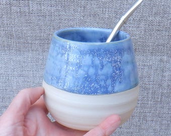May include: A ceramic mate cup featuring a rounded shape with a smooth, matte off-white base and a glossy, mottled blue upper section. A slender, metallic silver-toned bombilla straw is inserted into the opening of the vessel, which has a wide rim.