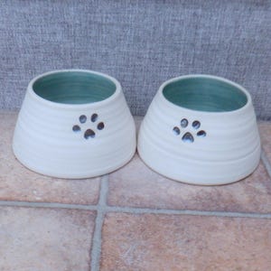 May include: Two white ceramic pet bowls with a teal blue interior and a paw print design.