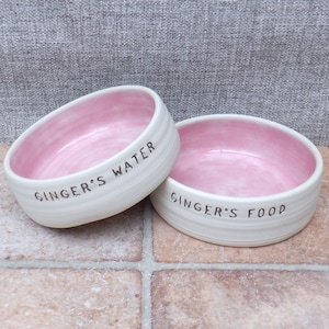 May include: Two white ceramic pet bowls with pink interiors. The bowls are personalised with the text "Ginger's Water" and "Ginger's Food".